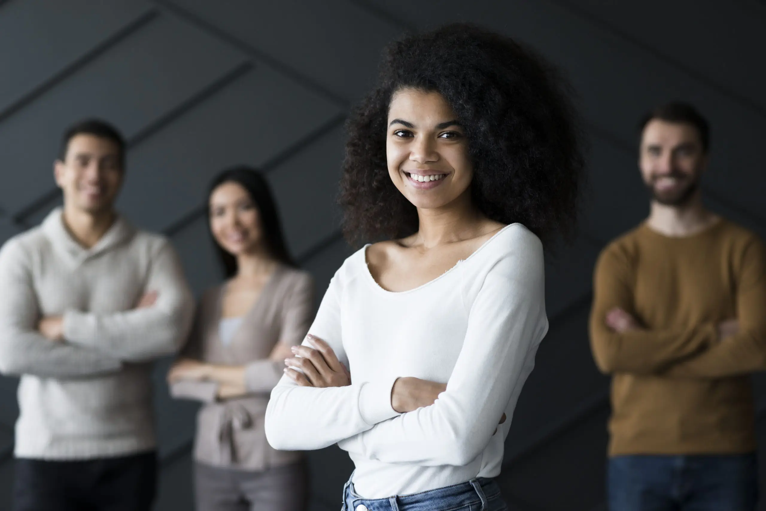 Grupo de pessoas numa empresa posam para uma foto de pé e braços cruzados sorrindo em frente a um fundo cinza opaco. À frente, a líder, uma jovem mulher negra de blusa branca. Ao fundo desfocado, três colaboradores, também jovens, brancos. A imagem ilustra um artigo de como o RH Digital traz vantagens para o setor.