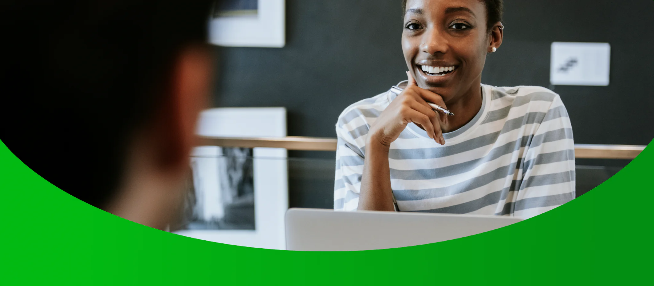 Jovem gestora de RH negra em seu escritório, senta-se à mesa com seu laptop sorridente com a mão ao queixo segurando uma caneta enquanto conversa com uma pessoa a sua frente de costas para a câmera ilustrando um diálogo sobre software para RH.